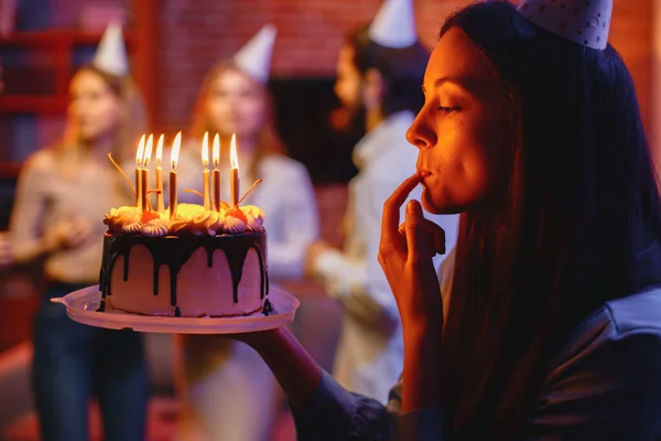 Woman in party cone holding a plate with birthday cake. Cake with cream and candles. Blurred silhouettes of people on a party behind.
