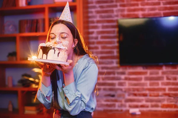 Young woman dips face in white cake with cream. Brunette girl wearing blue shirt and birthday cone hat. Birthday face cake smashing tradition concept.