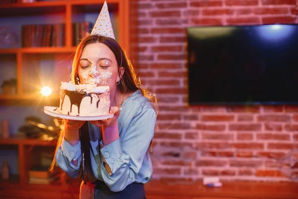 Young woman dips face in white cake with cream. Brunette girl wearing blue shirt and birthday cone hat. Birthday face cake smashing tradition concept.