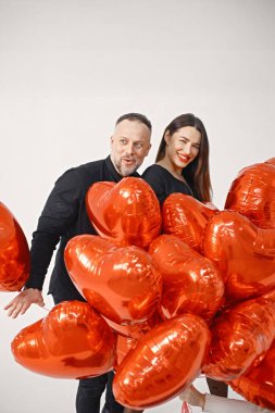 Yıldönümünde fotoğraf çektirmek için poz veren romantik bir çift. Adamın karısını kucakladığı ve elinde kalp şeklinde kırmızı balonlar tuttuğu bir stüdyo fotoğrafı. Esmer kadın ve siyah elbiseli sakallı adam.
