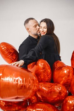 Yıldönümünde fotoğraf çektirmek için poz veren romantik bir çift. Adamın karısını kucakladığı ve elinde kalp şeklinde kırmızı balonlar tuttuğu bir stüdyo fotoğrafı. Esmer kadın ve siyah elbiseli sakallı adam.