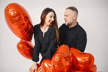 Yıldönümünde fotoğraf çektirmek için poz veren romantik bir çift. Adamın karısını kucakladığı ve elinde kalp şeklinde kırmızı balonlar tuttuğu bir stüdyo fotoğrafı. Esmer kadın ve siyah elbiseli sakallı adam.