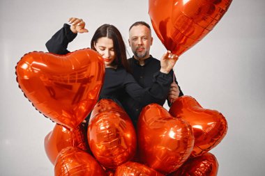 Yıldönümünde fotoğraf çektirmek için poz veren romantik bir çift. Adamın karısını kucakladığı ve elinde kalp şeklinde kırmızı balonlar tuttuğu bir stüdyo fotoğrafı. Esmer kadın ve siyah elbiseli sakallı adam.