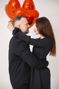 Yıldönümünde fotoğraf çektirmek için poz veren romantik bir çift. Adamın karısını kucakladığı ve elinde kalp şeklinde kırmızı balonlar tuttuğu bir stüdyo fotoğrafı. Esmer kadın ve siyah elbiseli sakallı adam.