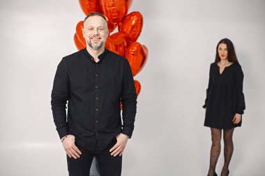 Önde duran bir adam fotoğraf için poz veriyor. Bir grup kalp şeklinde kırmızı balonun arkasında duran bir kadının uzun çekim stüdyosu. Esmer kadın ve siyah elbiseli sakallı adam..