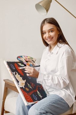 Tatmin olmuş kadın sanatçı elinde yeni bir resim ve elinde boya paleti ile poz veriyor. Kot pantolon ve beyaz gömlek giyen bir kadın. Beyaz duvarlı bir sandalyede oturan esmer kız..