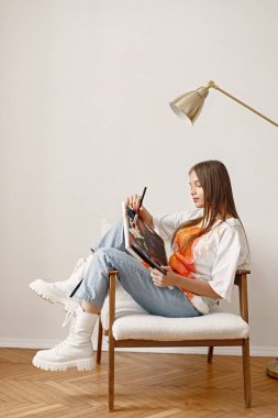 Fırça tutarken ve resim yaparken poz veren genç bir kadın sanatçı. Beyaz tişört ve kot pantolon giyen bir kadın. Beyaz duvarlı bir sandalyede oturan esmer kız..