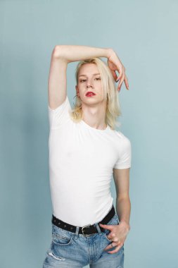 Makyajlı genç sarışın bir adam fotoğraf için poz veriyor. Beyaz tişörtlü ve kırmızı dudaklı bir adam. Mavi arkaplanda izole edilmiş bir adam.