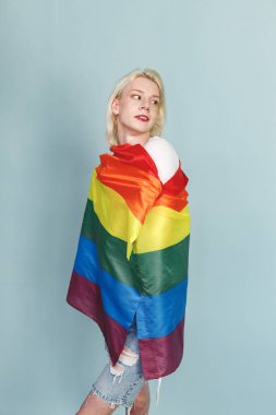 Yüzünde makyaj ve gökkuşağı bayrağı olan genç sarışın bir adam fotoğraf için poz veriyor. Beyaz tişörtlü ve kırmızı dudaklı bir adam. Mavi arkaplanda izole edilmiş bir adam.