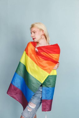 Yüzünde makyaj ve gökkuşağı bayrağı olan genç sarışın bir adam fotoğraf için poz veriyor. Beyaz tişörtlü ve kırmızı dudaklı bir adam. Mavi arkaplanda izole edilmiş bir adam.