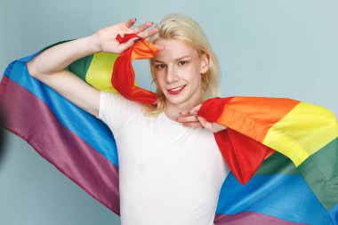 Yüzünde makyaj ve gökkuşağı bayrağı olan genç sarışın bir adam fotoğraf için poz veriyor. Beyaz tişörtlü ve kırmızı dudaklı bir adam. Mavi arkaplanda izole edilmiş bir adam.