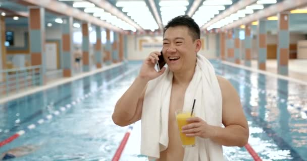 スイマーは屋内プールでスマートフォンで話します 男の韓国国籍プールに立って電話で話してジュースを飲む — ストック動画