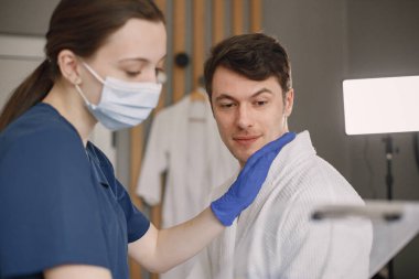 Adam kozmetolog kabinesinde oturuyor ve kozmetologla konuşuyor. Mavi tıbbi kostüm ve yüz maskesi takan kadın kozmetik uzmanı. Beyaz bornoz giyen adam..