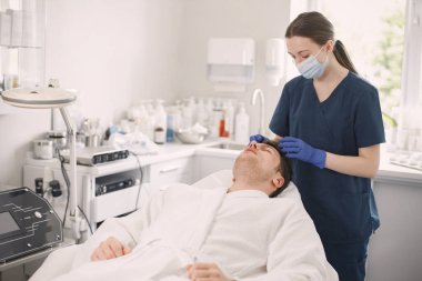 Adam kozmetolog kabinesinde yatıyor ve yüz derisi için bir prosedürü var. Mavi tıbbi kostüm ve yüz maskesi takan kadın kozmetik uzmanı. Beyaz bornoz giyen adam..