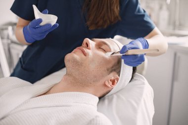 Adam kozmetolog kabinesinde yatıyor ve yüz derisi için bir prosedürü var. Mavi tıbbi kostüm giyen tanınmamış bir kadın kozmetik uzmanı. Erkek yüzüne maske takan bir kadın..