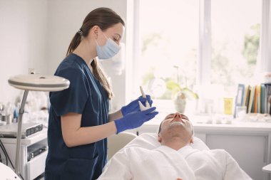 Adam kozmetolog kabinesinde yatıyor ve yüz derisi için bir prosedürü var. Mavi tıbbi kostüm ve yüz maskesi takan kadın kozmetik uzmanı. Erkek yüzüne maske takan bir kadın..