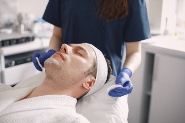 Adam kozmetolog kabinesinde yatıyor ve yüz derisi için bir prosedürü var. Mavi tıbbi kostüm giyen tanınmamış bir kadın kozmetik uzmanı. Erkek yüzüne maske takan bir kadın..