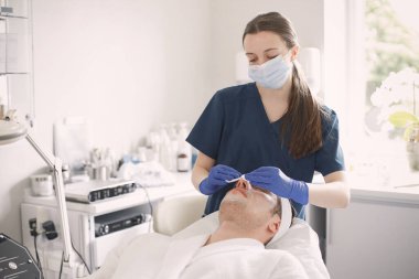 Kozmetolog kabinesinde yatan ve yüz derisi için bir prosedürü olan bir adam. Mavi tıbbi kostüm giyen bir kadın kozmetikçi. Erkeklerin yüzünü pamukla silen kadın..