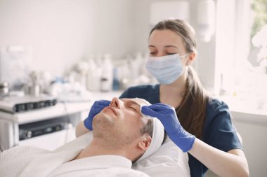 Kozmetolog kabinesinde yatan ve yüz derisi için bir prosedürü olan bir adam. Mavi tıbbi kostüm giyen bir kadın kozmetikçi. Erkeklerin yüzünü pamukla silen kadın..