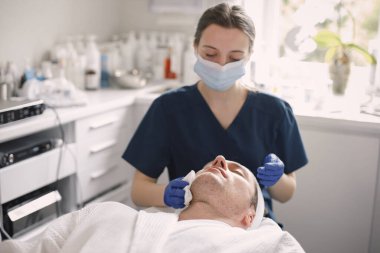 Kozmetolog kabinesinde yatan ve yüz derisi için bir prosedürü olan bir adam. Mavi tıbbi kostüm giyen bir kadın kozmetikçi. Kadın, erkeğin yüzünü bir bezle siliyor..