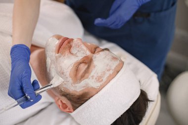 Adam kozmetolog kabinesinde yatıyor ve yüz derisi için bir prosedürü var. Mavi tıbbi kostüm giyen tanınmamış bir kadın kozmetik uzmanı. Erkek yüzüne maske takan bir kadın..