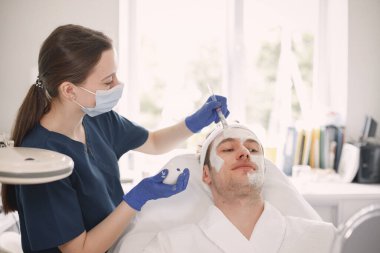 Adam kozmetolog kabinesinde yatıyor ve yüz derisi için bir prosedürü var. Mavi tıbbi kostüm ve yüz maskesi takan kadın kozmetik uzmanı. Beyaz bornoz giyen adam..