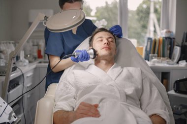 Kozmetolog kabinesinde yatan genç bir adam ve yüz derisi için bir çürüme prosedürü var. Mavi tıbbi kostüm ve yüz maskesi takan kadın kozmetik uzmanı. Beyaz bornoz giyen adam..