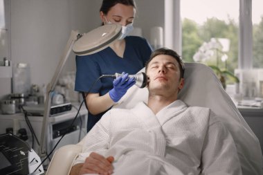 Kozmetolog kabinesinde yatan genç bir adam ve yüz derisi için bir çürüme prosedürü var. Mavi tıbbi kostüm ve yüz maskesi takan kadın kozmetik uzmanı. Beyaz bornoz giyen adam..