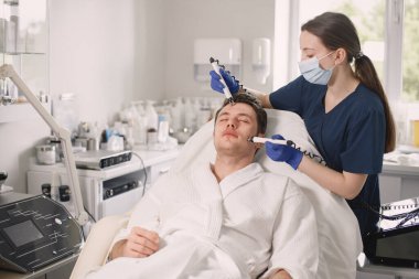 Kozmetolog kabinesinde yatan genç bir adam ve yüz derisi için bir çürüme prosedürü var. Mavi tıbbi kostüm ve yüz maskesi takan kadın kozmetik uzmanı. Beyaz bornoz giyen adam..