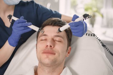 Kozmetik bilimci kabinesinde yatan ve yüz derisi için çürüme prosedürü olan genç bir adama yakın çekim. Mavi tıbbi kostüm ve eldiven giyen kadın kozmetik uzmanı. Beyaz bornoz giyen adam..