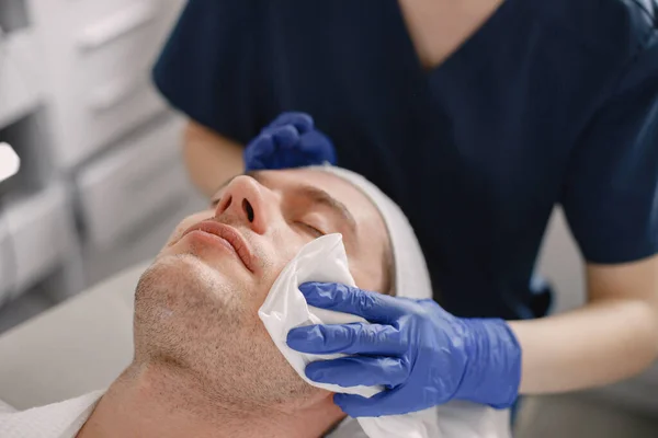 Man laying in cosmetologist cabinet and has a procedure for face skin.Female cosmetologist wearing blue medical costume. Woman wiping mans face with a cloth.