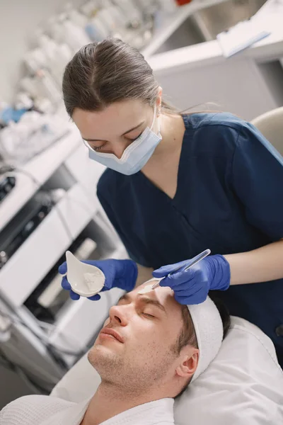 Man laying in cosmetologist cabinet and has a procedure for face skin. Female cosmetologist wearing blue medical costume and face mask. Woman applying a mask on mans face.