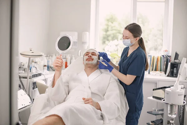 Young man laying in cosmetologist cabinet and has a procedure for face skin. Female cosmetologist wearing blue medical costume and face mask. Man wearing white bathrobe and looking in the mirror.