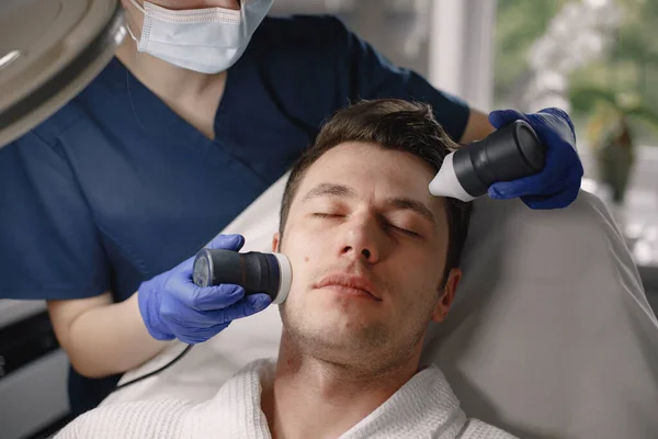 Young man laying in cosmetologist cabinet and has a cavitation procedure for face skin. Female cosmetologist wearing blue medical costume and face mask. Man wearing white bathrobe.