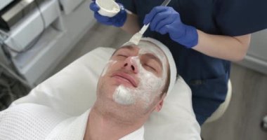 Kozmetik uzmanı spa salonundaki erkeklerin yüzüne kilden maske takıyor. Güzellik uzmanı, güzellik salonunda yakışıklı bir adam için tedavi görüyor..
