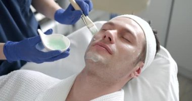 Cosmetologist applying nourishing mask on man client face in beauty clinic, closeup.