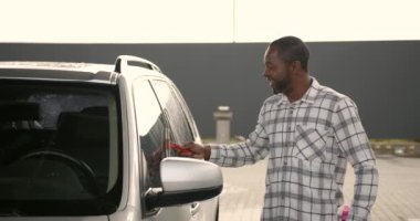 Car detailing. Black man wiping water from car window with squeegee rubber by hand.