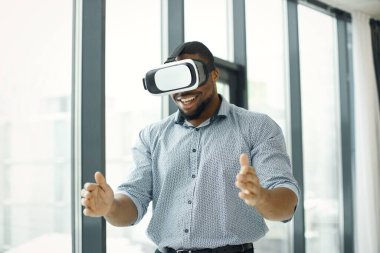 Black business man standing in office near big windows. Bearded man wearing virtual reality glasses. Man wearing blue shirt.
