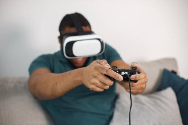 Black man in vr glasses, playing video games with virtual reality headset. Man wearing blue t-shirt. Man in virtual reality glasses sitting at home on a coach.