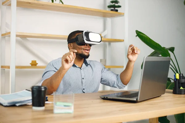 Black business man sitting in office with a laptop. Bearded man using virtual reality glases. Man wearing blue shirt.