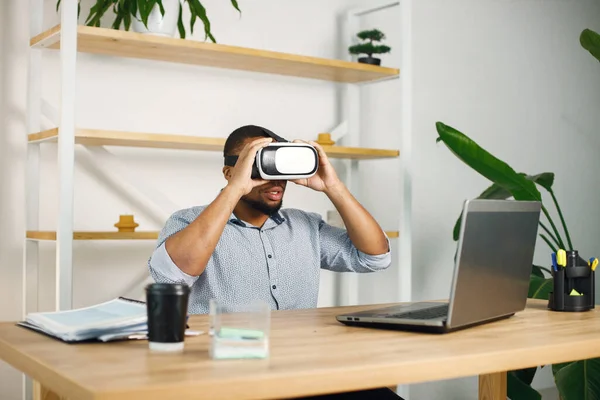 Black business man sitting in office with a laptop. Bearded man using virtual reality glases. Man wearing blue shirt.