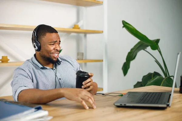 Black business man sitting in office with a laptop. Bearded man in earphones has a video call. Man wearing blue shirt and holding takeaway coffee..