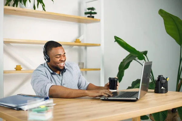 Black business man sitting in office with a laptop. Bearded man in earphones has a video call. Man wearing blue shirt and holding takeaway coffee..