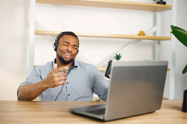 Black business man sitting in office with a laptop. Bearded man in earphones has a video call. Man wearing blue shirt and holding takeaway coffee.