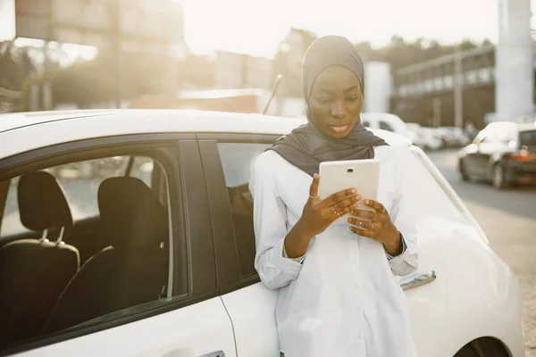 African muslim woman leaning on her car and holding a digital tablet. Working remotely or sharing info. Technologies in our life.