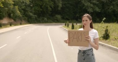 Beautiful female hitchhiker with cardboard standing on highway and catching passing car. Hitchhiking concept.