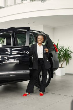 Young and cheerful woman enjoying new car while standing behinde. Black woman buy a new car. Girl wearing black costume.