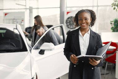 Professional car dealer standing at auto showroom and holding clipboard. Two customers behind looking on a car. Black woman wearing a black costume and white shirt.