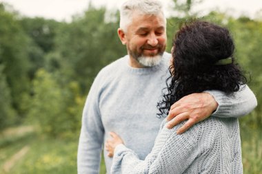 Close up romantic couple standing in autumn park and hugging in daytime. Man and woman wearing blue sweaters. Woman is brunette and man is gray.