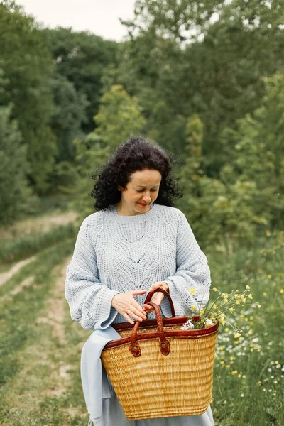 Senior woman walking through the park alone. Woman wearing blue sweater and skirt. Brunette woman holding a bag with a flowers.